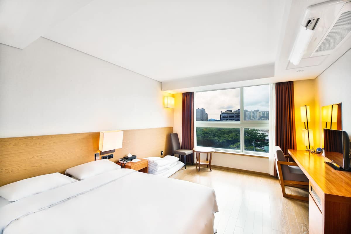 A hotel room with a large bed, a desk with a computer, a chair, and a window with a city view.
