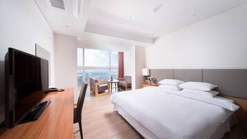 A modern hotel room with a large bed, a desk with a computer, a TV, a chair, and a small table.