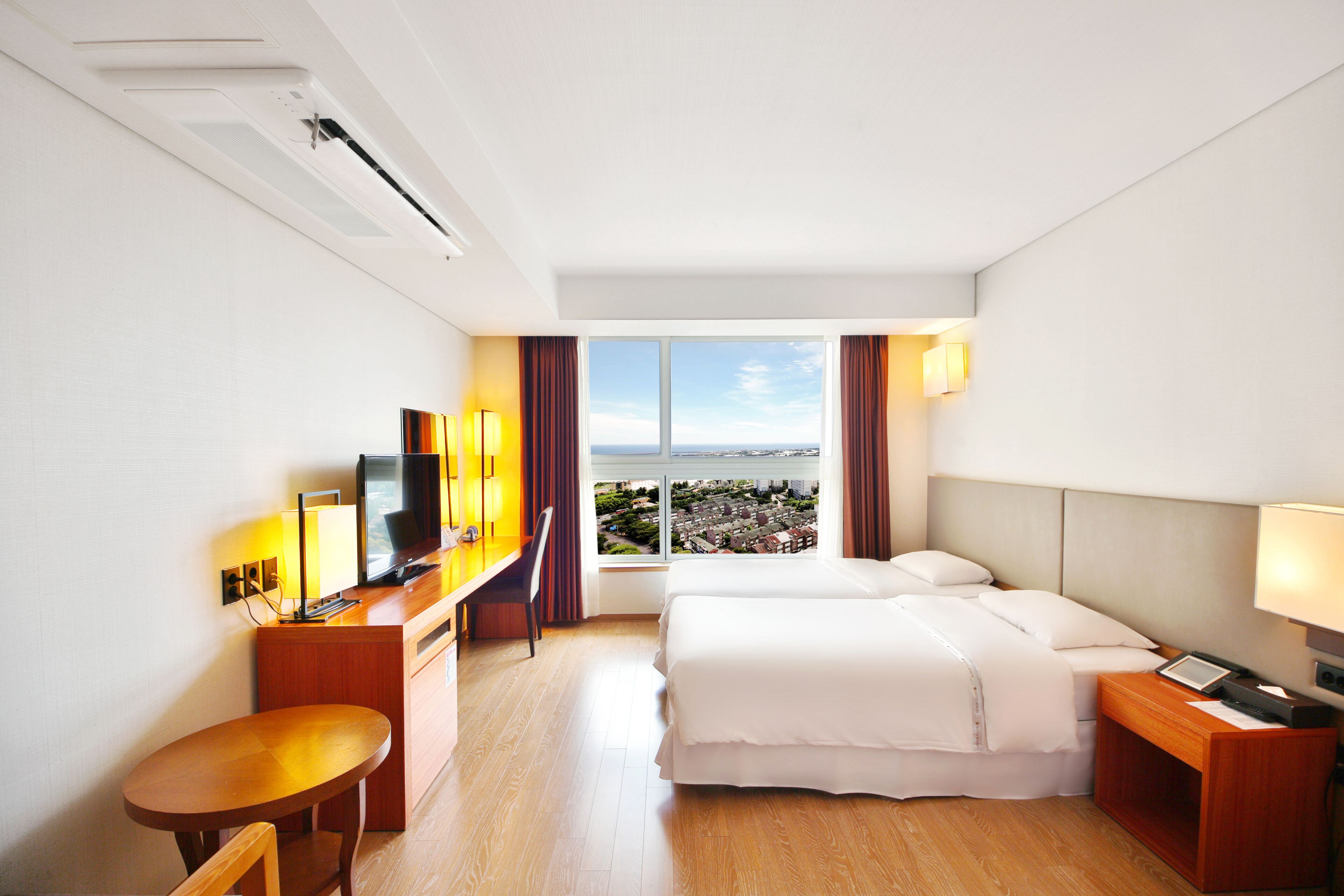 A hotel room with a bed, a desk with a TV, a chair, a small table, and a window with a city view.