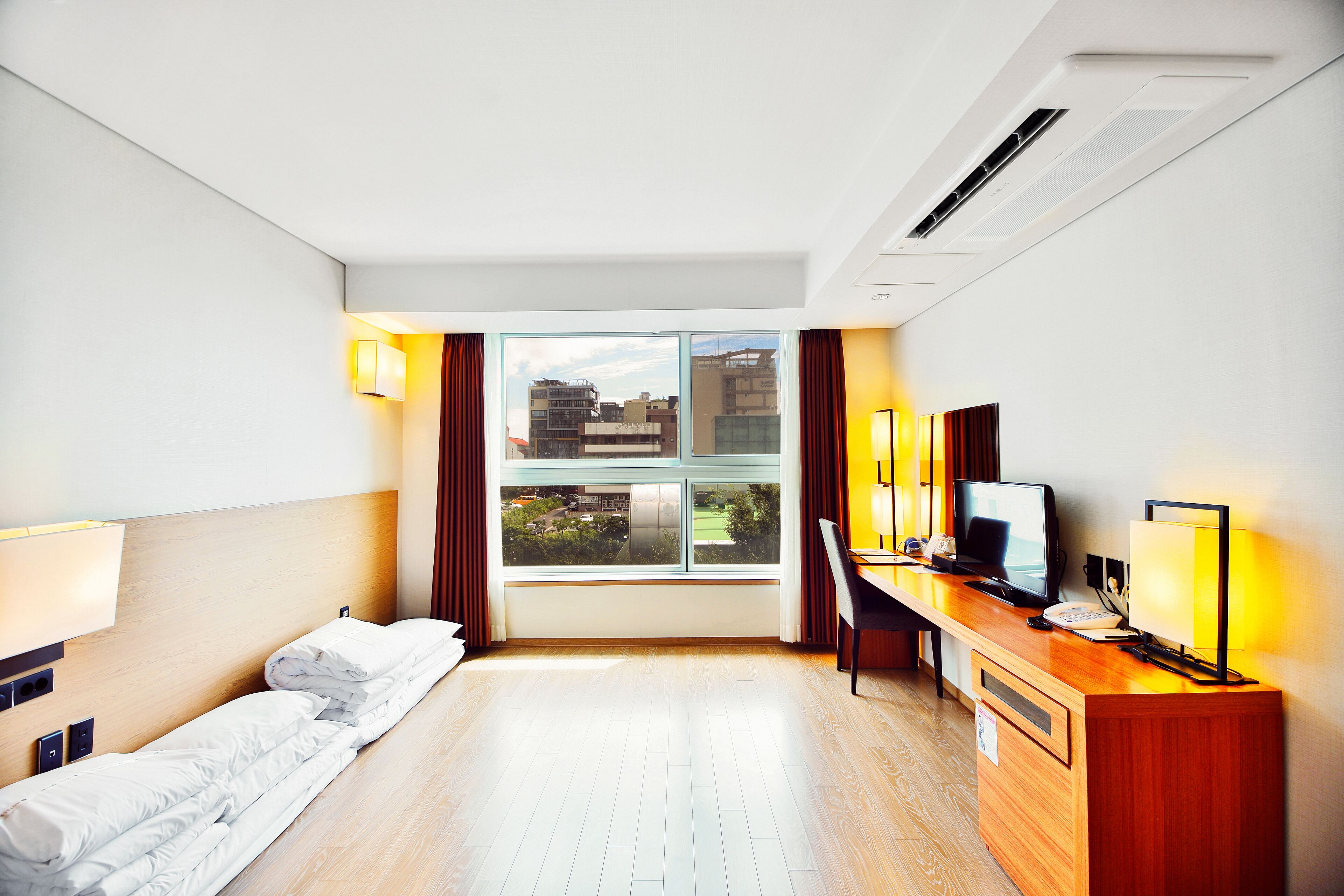 A hotel room with a bed, desk, computer, and a window with a view of buildings.