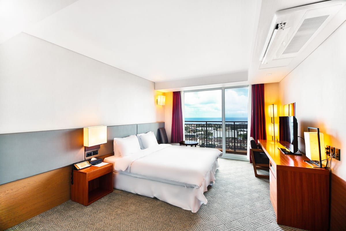 A hotel room with a bed, desk, and a view of the city through large windows.