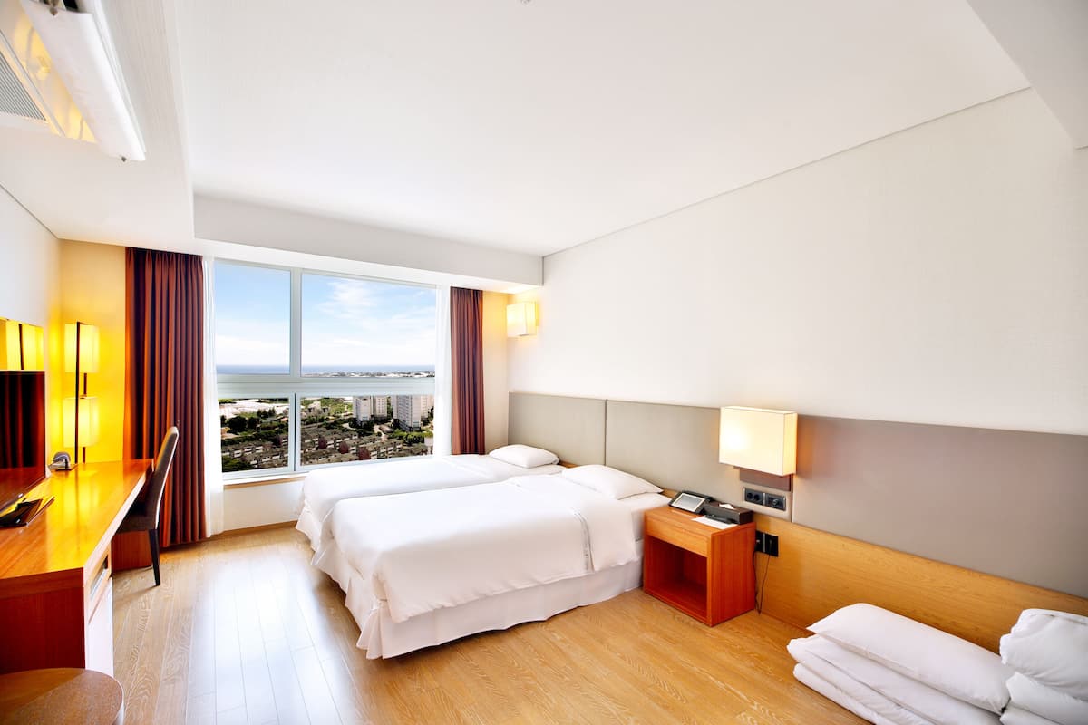 A hotel room with two beds, a desk, a chair, a nightstand, and a view of the city through large windows.