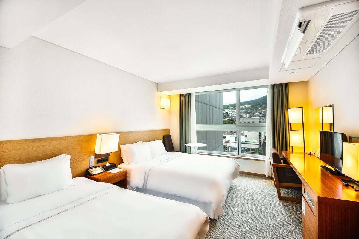 A hotel room with two beds, a desk with a TV, and a window with a city view.