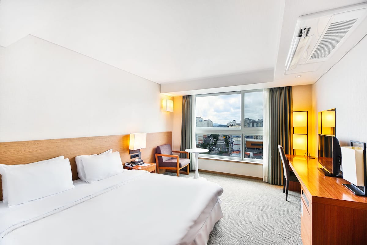 A hotel room with a large bed, a desk with a chair, a television, and a window with a city view.