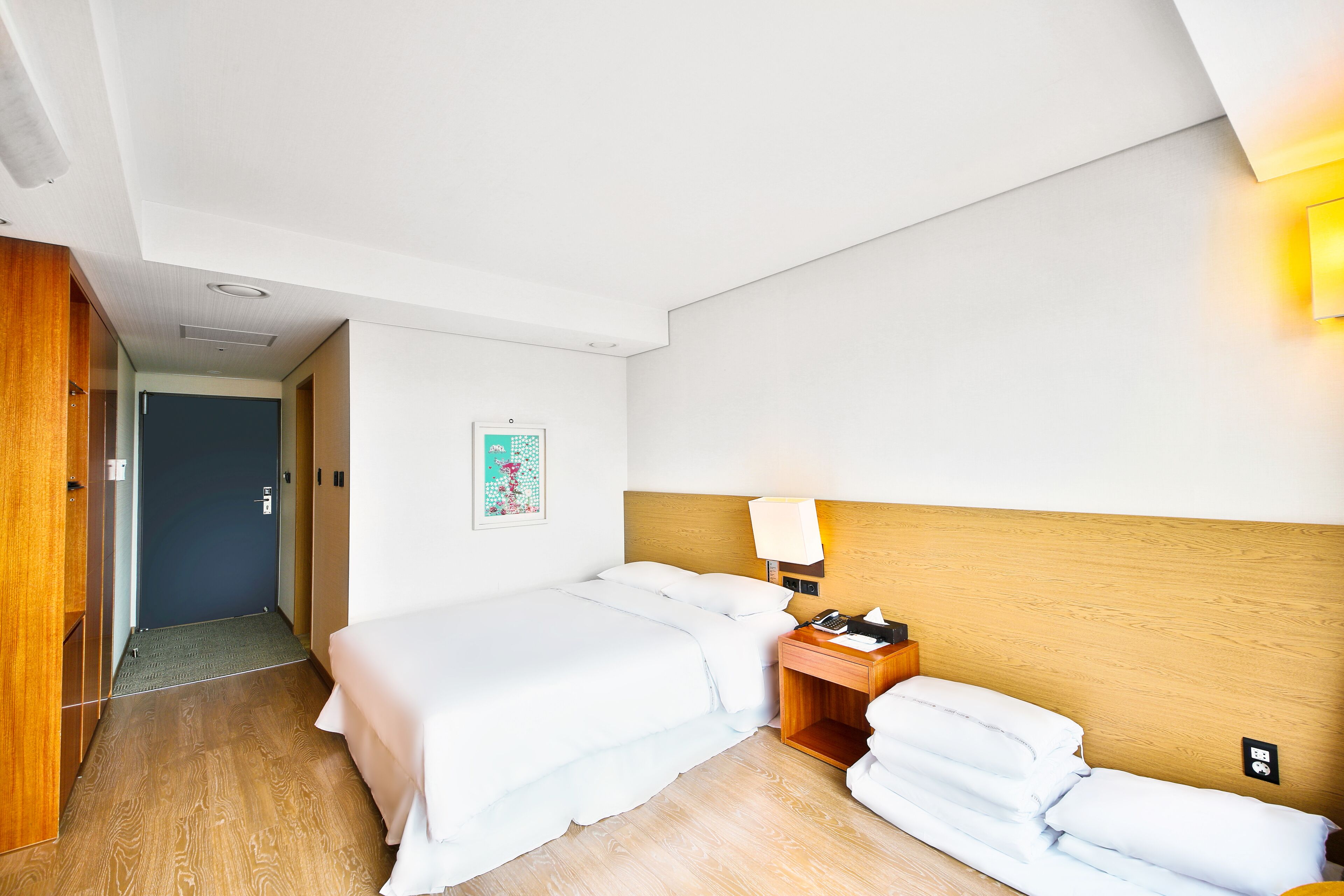 Habitación de hotel con piso de madera, cama con ropa blanca, mesita de noche con lámpara y puerta que da a otra habitación.