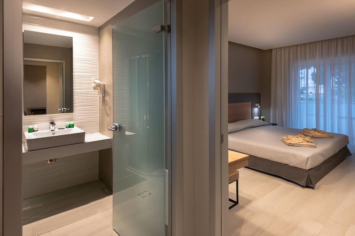 Een moderne hotelkamer met een douchecabine van glas, een nachtkastje met een lamp, een bed met handdoeken en een groot raam met gordijnen.