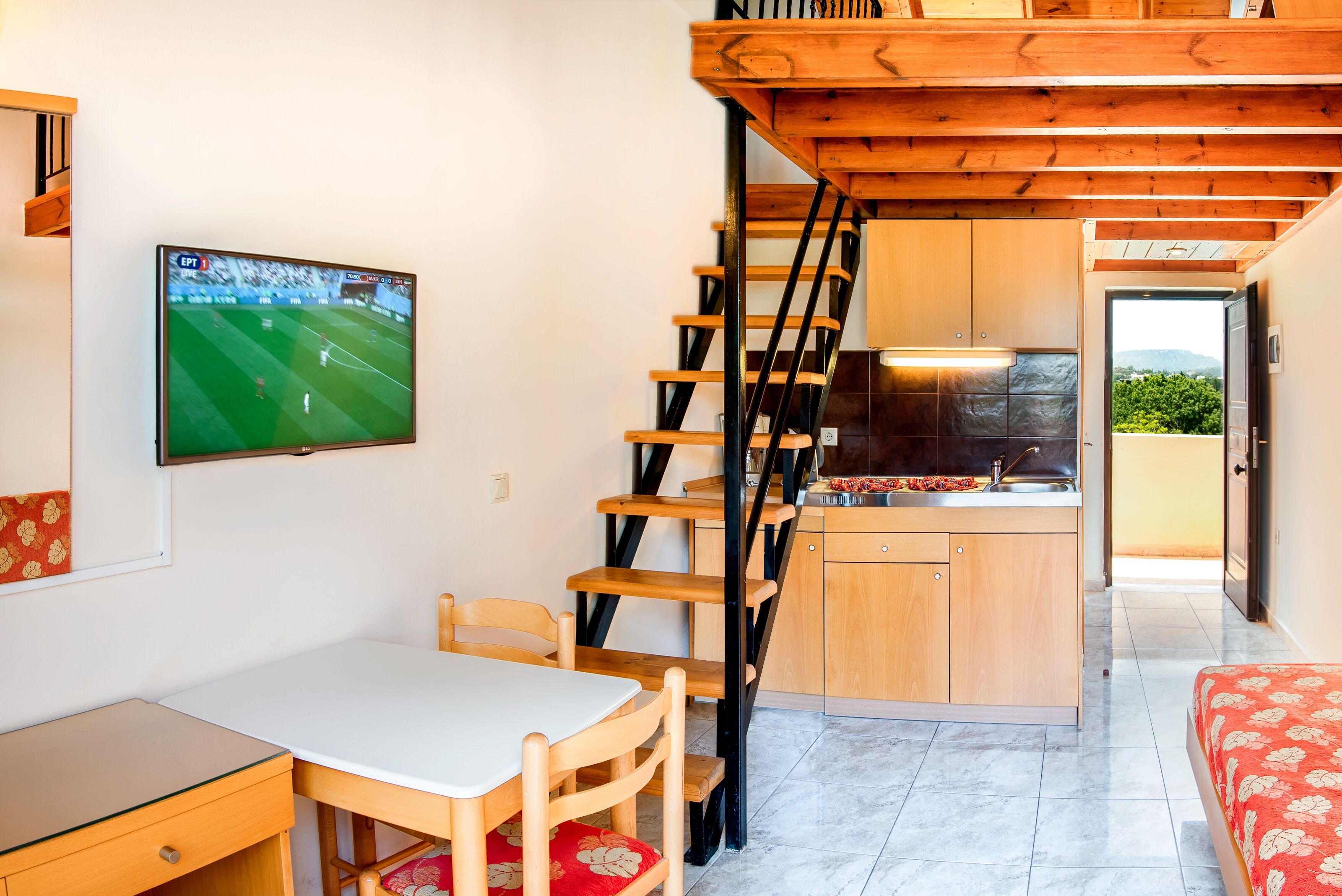 A compact room with a dining table, chairs, a TV mounted on the wall, a kitchen area with a sink and cabinets, and a staircase.