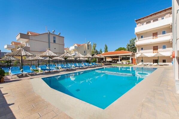 Una piscina con agua azul, rodeada de sillones de baño y sombrillas, junto a un edificio con balcones.
