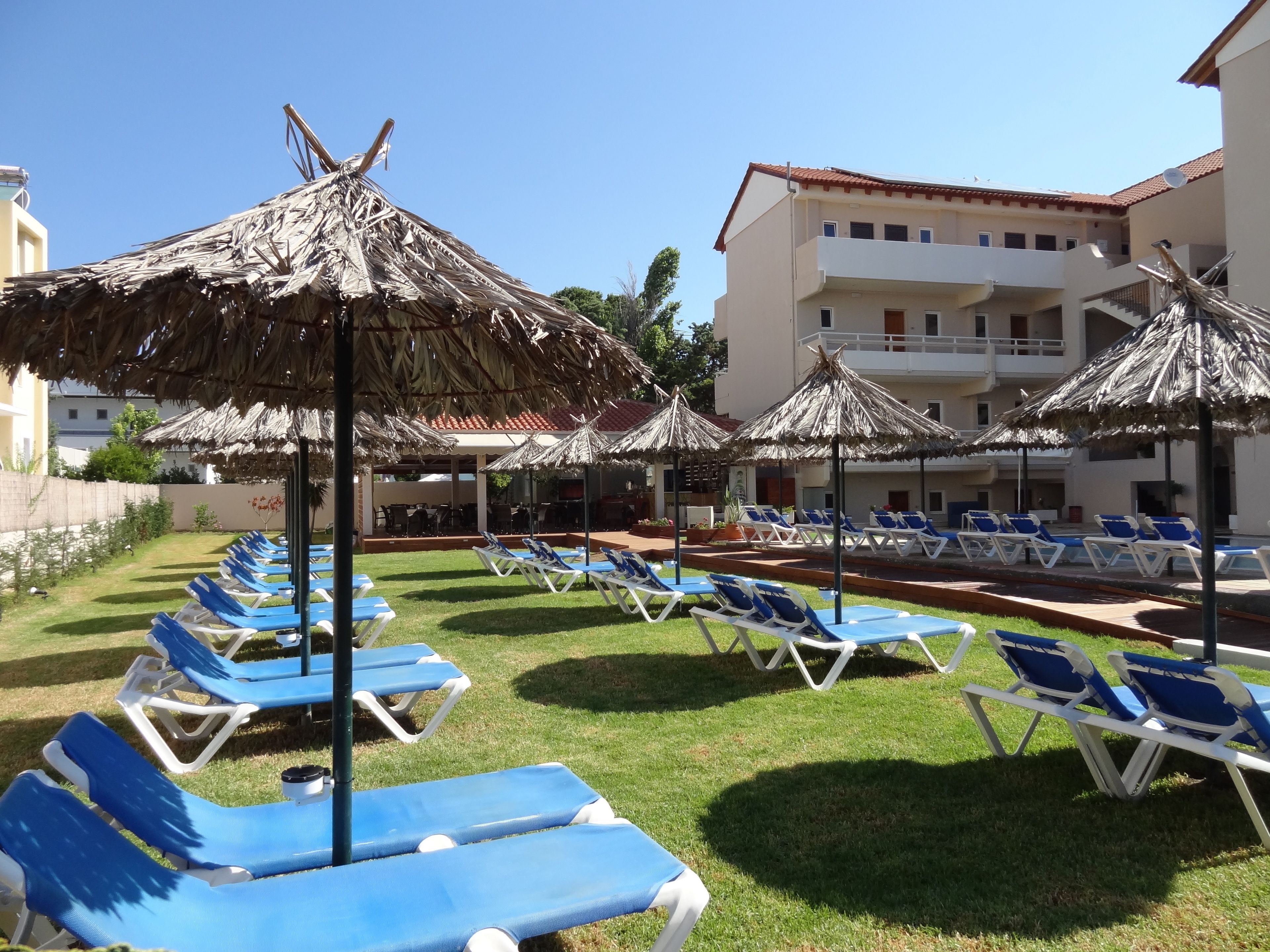 A poolside area with blue lounge chairs and thatched umbrellas, surrounded by a building with balconies.