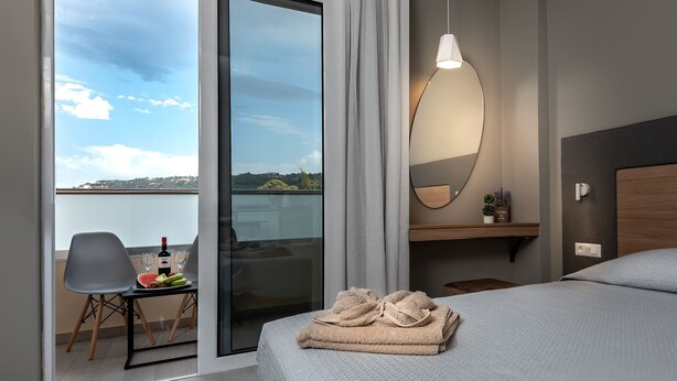 A hotel room with a bed, a chair, a table with a bottle of wine and fruit, a view of the sky and landscape, and a mirror on the wall. - Sunday Hotel (Rhodes)