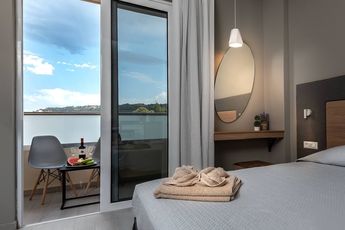 Een hotelkamer met een bed, een stoel, een tafel met wijn en fruit, uitzicht op de lucht en het landschap, en een spiegel aan de muur.