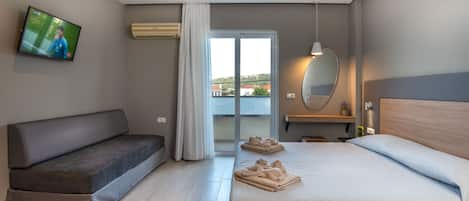 A modern hotel room with a sofa, a bed with towels, a TV, a sliding glass door, and a view of buildings outside.