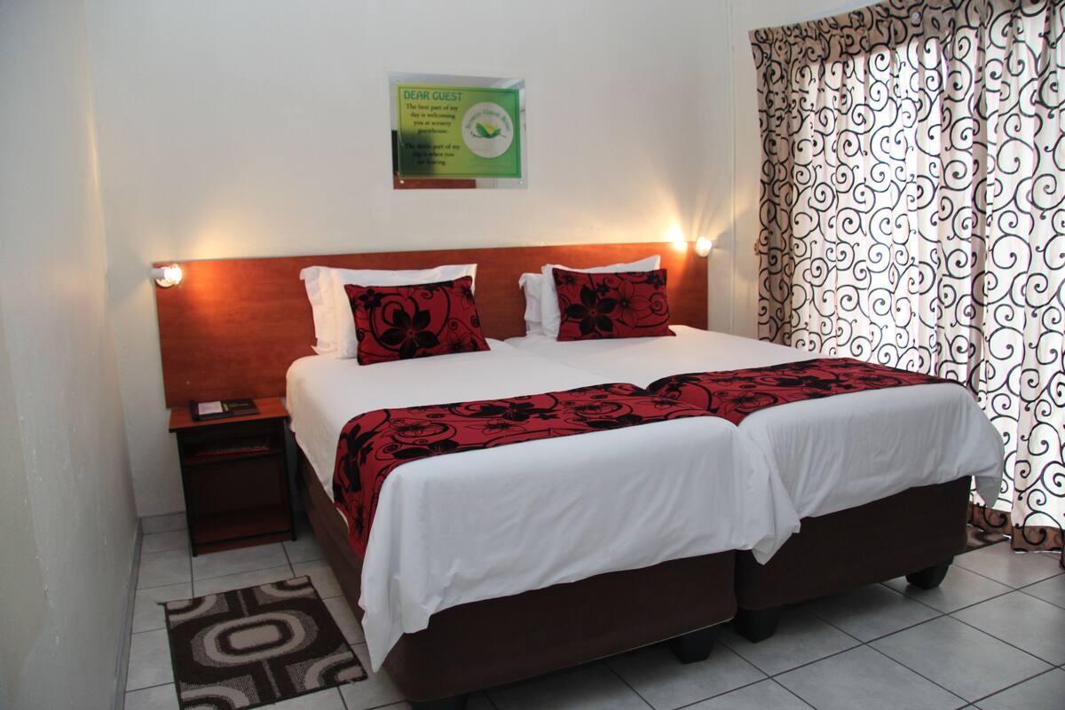 A double bed with a white and red bedspread, two red pillows, a wooden headboard, a nightstand with a lamp, and a patterned curtain.