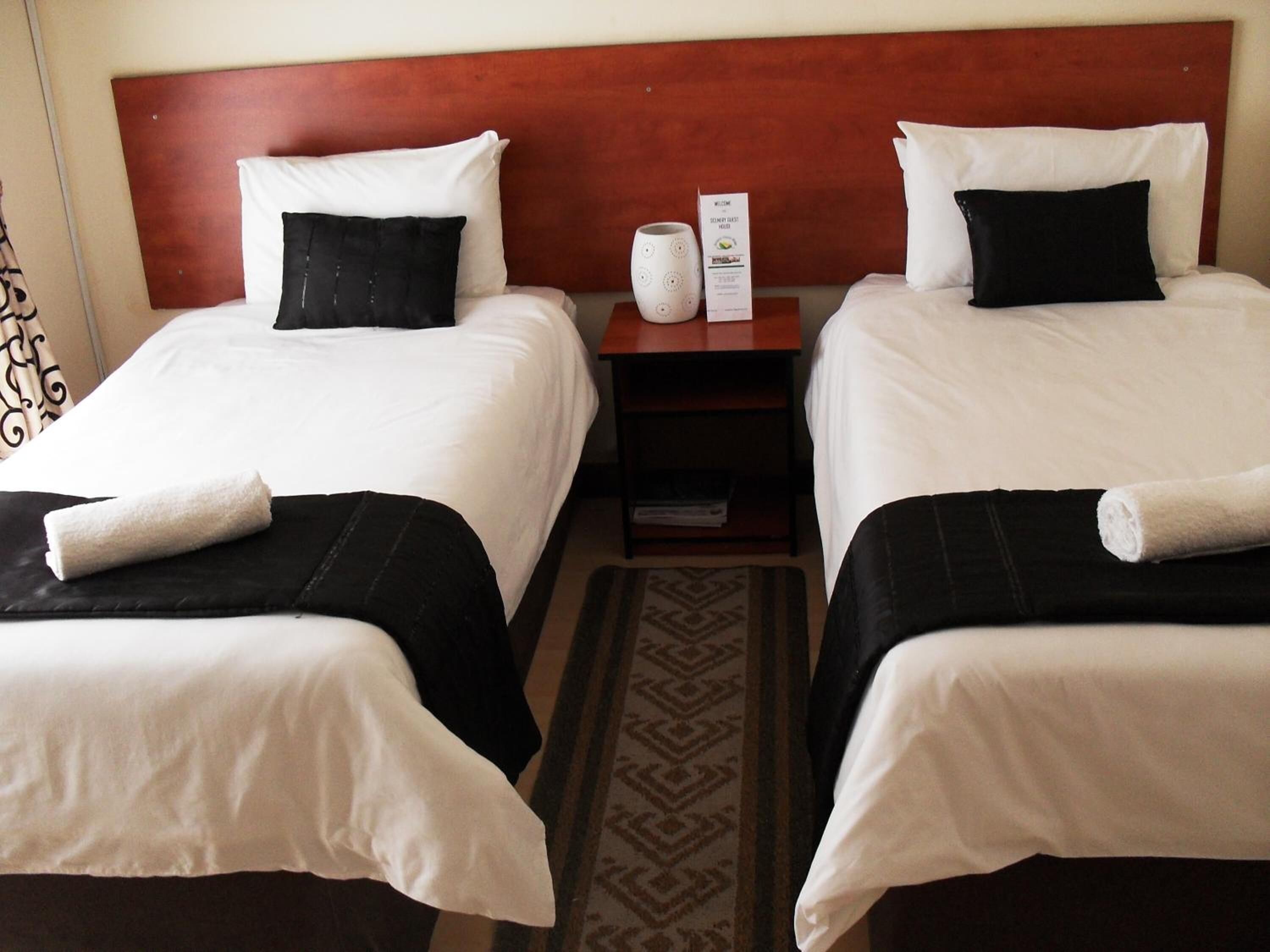 A hotel room with two beds, each with white linens and black pillows, a wooden headboard, a nightstand with a white vase and a welcome card, and a patterned rug.