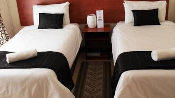 A hotel room with two beds, each with white linens and black pillows, a wooden headboard, a nightstand with a white vase and a welcome card, and a patterned rug.