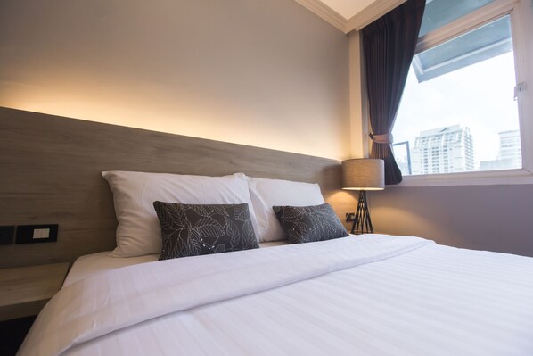 A hotel room with a large bed, two decorative pillows, a bedside lamp, and a view of the city through the window.
