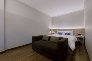A hotel room with a brown sofa, a bed with white bedding, a bedside table with a lamp, and a wooden floor.