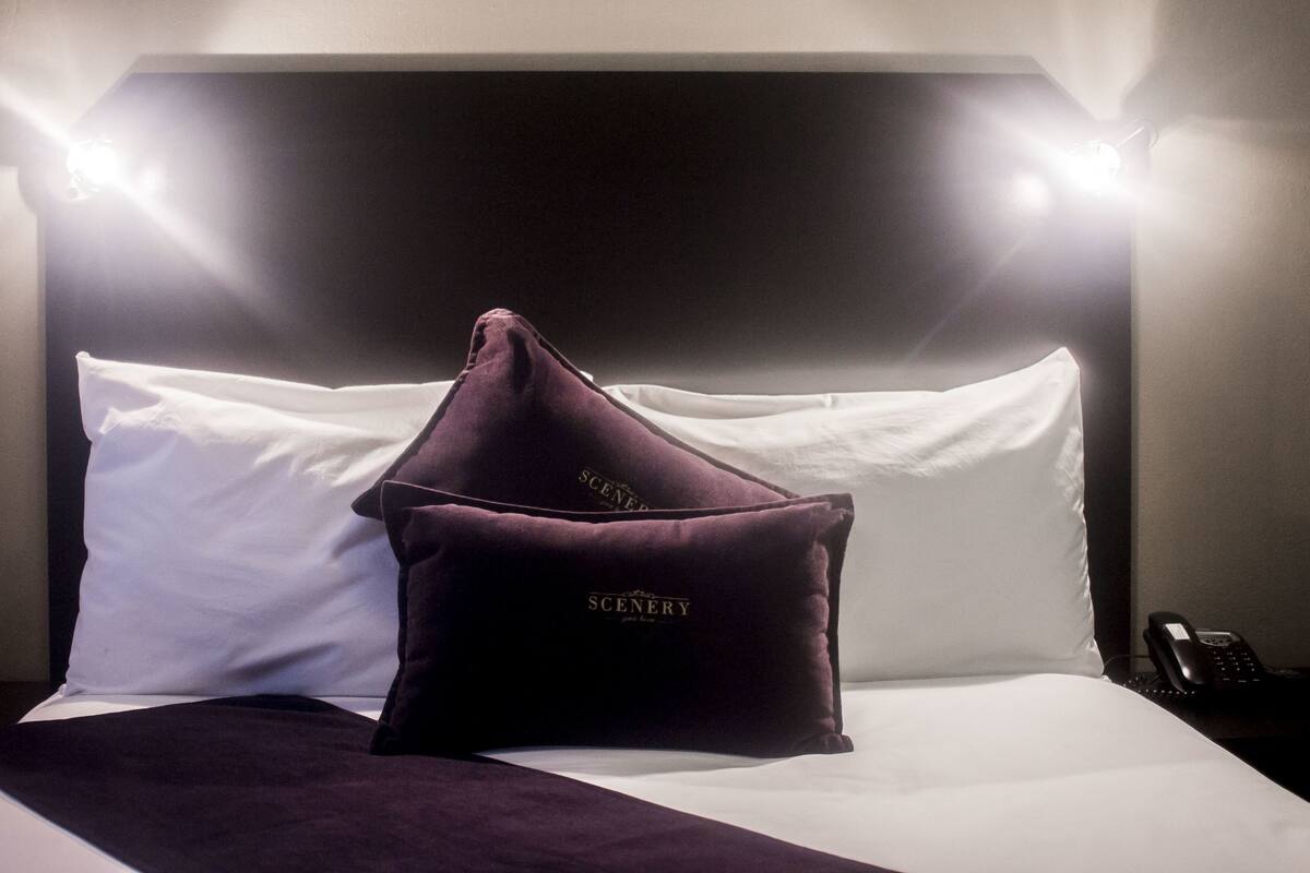 A hotel room with a neatly made bed, a headboard with two lights, and a pillow with the word "SCENERY" on it.