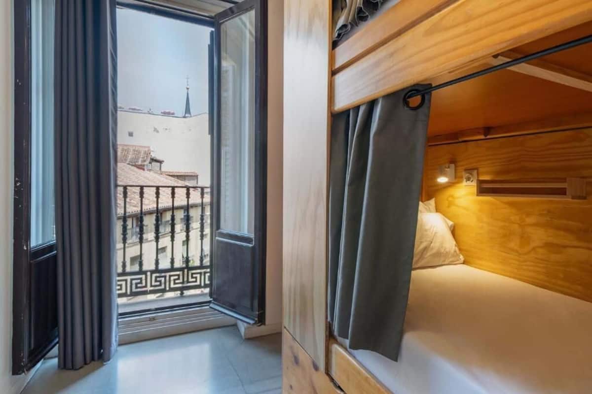 Una habitación con cama, ventana con cortinas y balcón con barandilla.