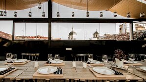 A restaurant with wooden tables set for dining, large windows offering a view of buildings, and hanging light fixtures. - The Hat Madrid (Madrid)
