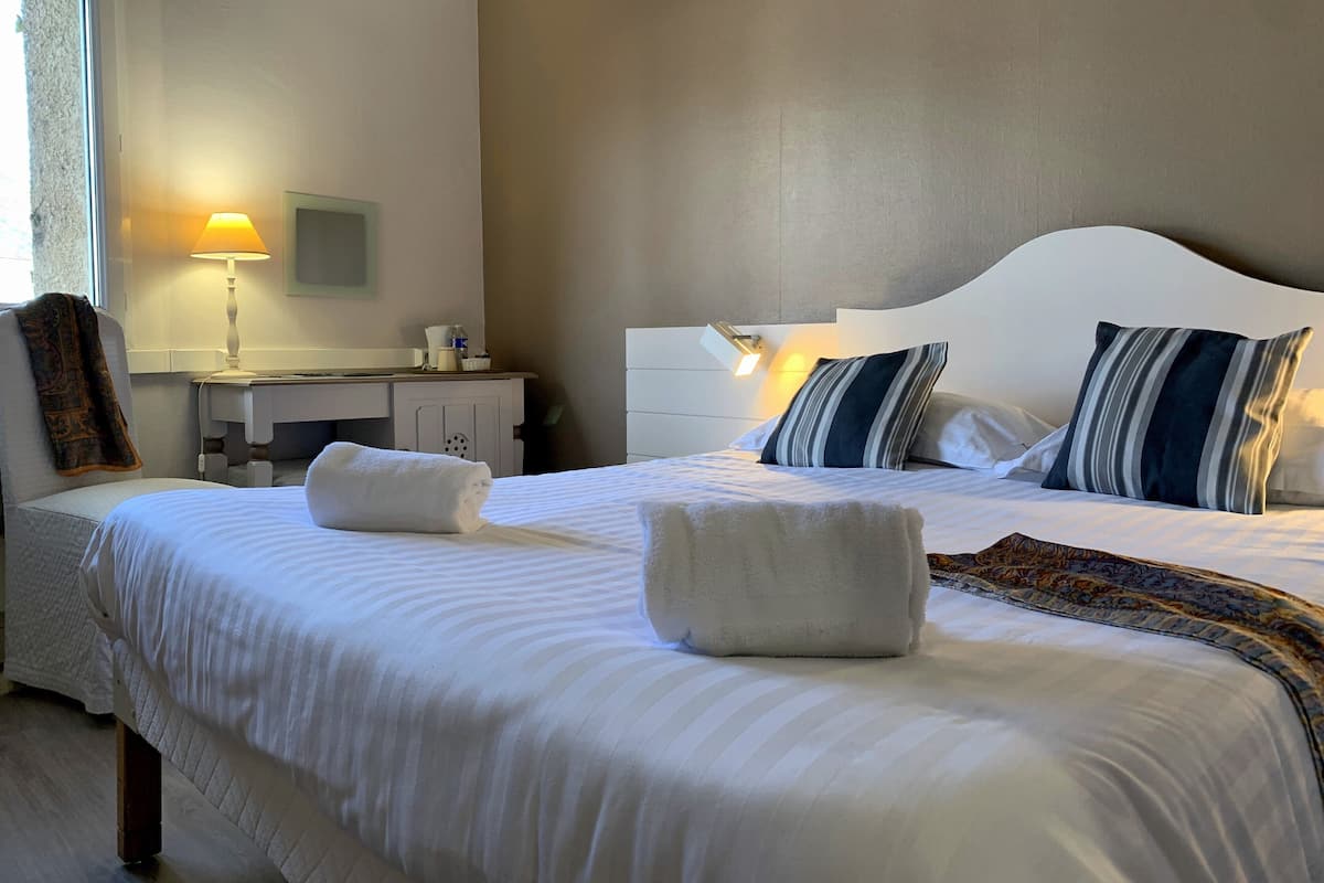Un lit bien fait, avec du linge de lit blanc et deux serviettes, une table de chevet avec une lampe, une petite armoire et un luminaire fixé au mur.