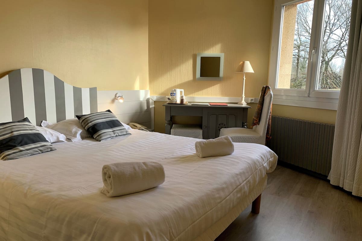 Une chambre d’hôtel avec un grand lit, des tables de chevet, un bureau, une chaise, une lampe, une télévision et une fenêtre donnant sur des arbres.