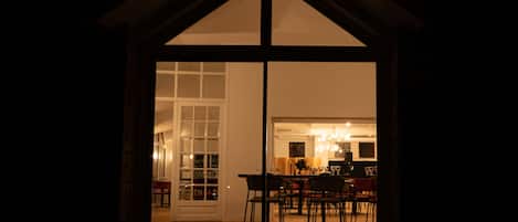 A view through a large window of a dining area with a table and chairs, a chandelier, and a kitchen in the background.