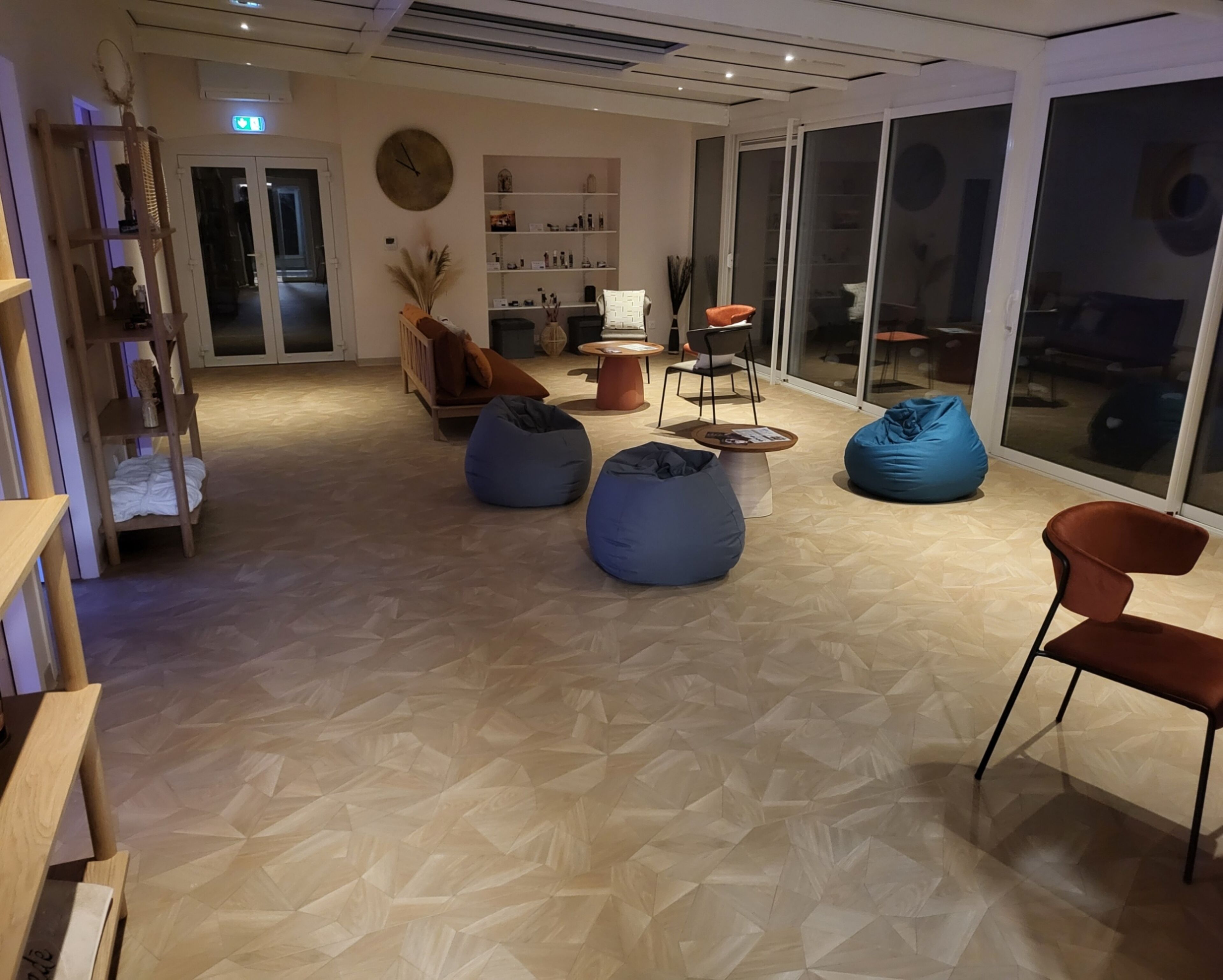 Un interior moderno con suelo de diseño geométrico, una estantería de madera, un reloj redondo y un área de descanso con pufs y sillas.