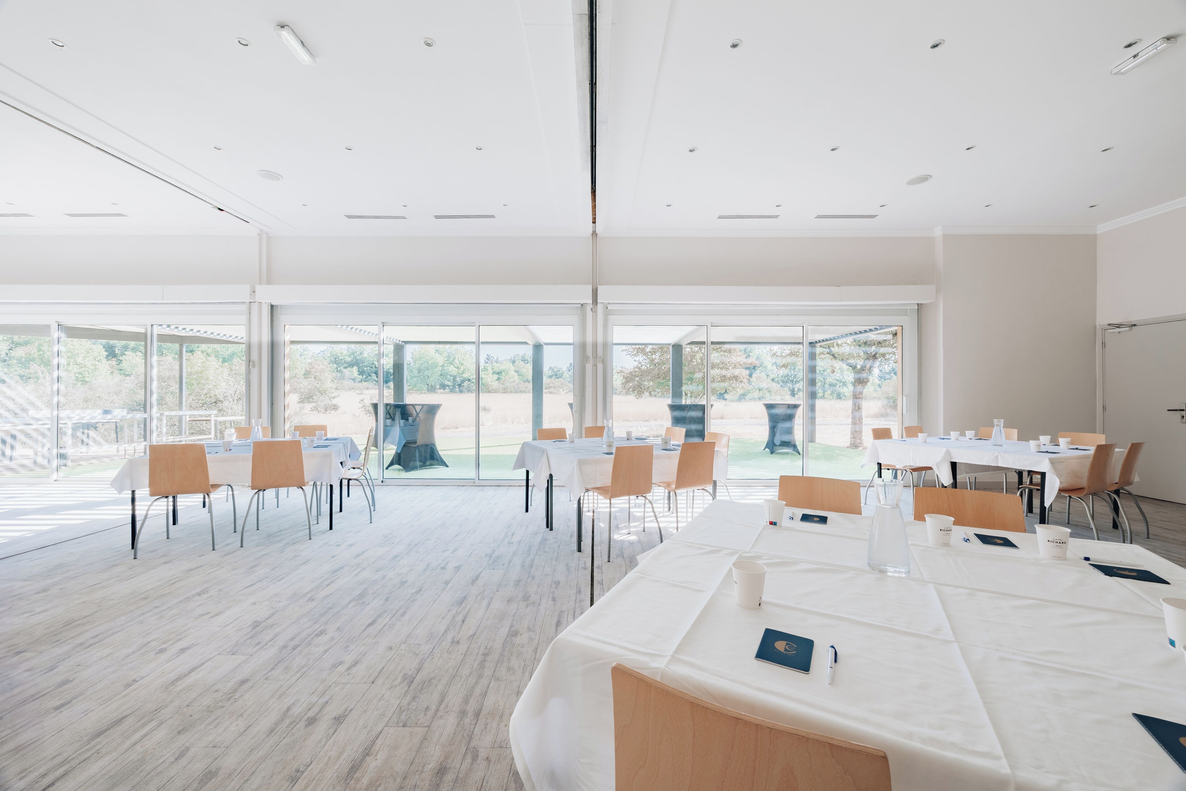 Un salón de conferencias amplio con mesas dispuestas para reuniones, amplias ventanas que ofrecen vistas al exterior y un techo moderno con iluminación empotrada.