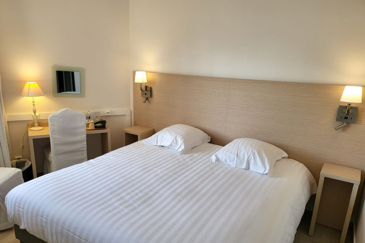 Un lit bien fait, avec du linge de lit blanc, deux tables de chevet avec des lampes, une chaise et un téléviseur fixé au mur.