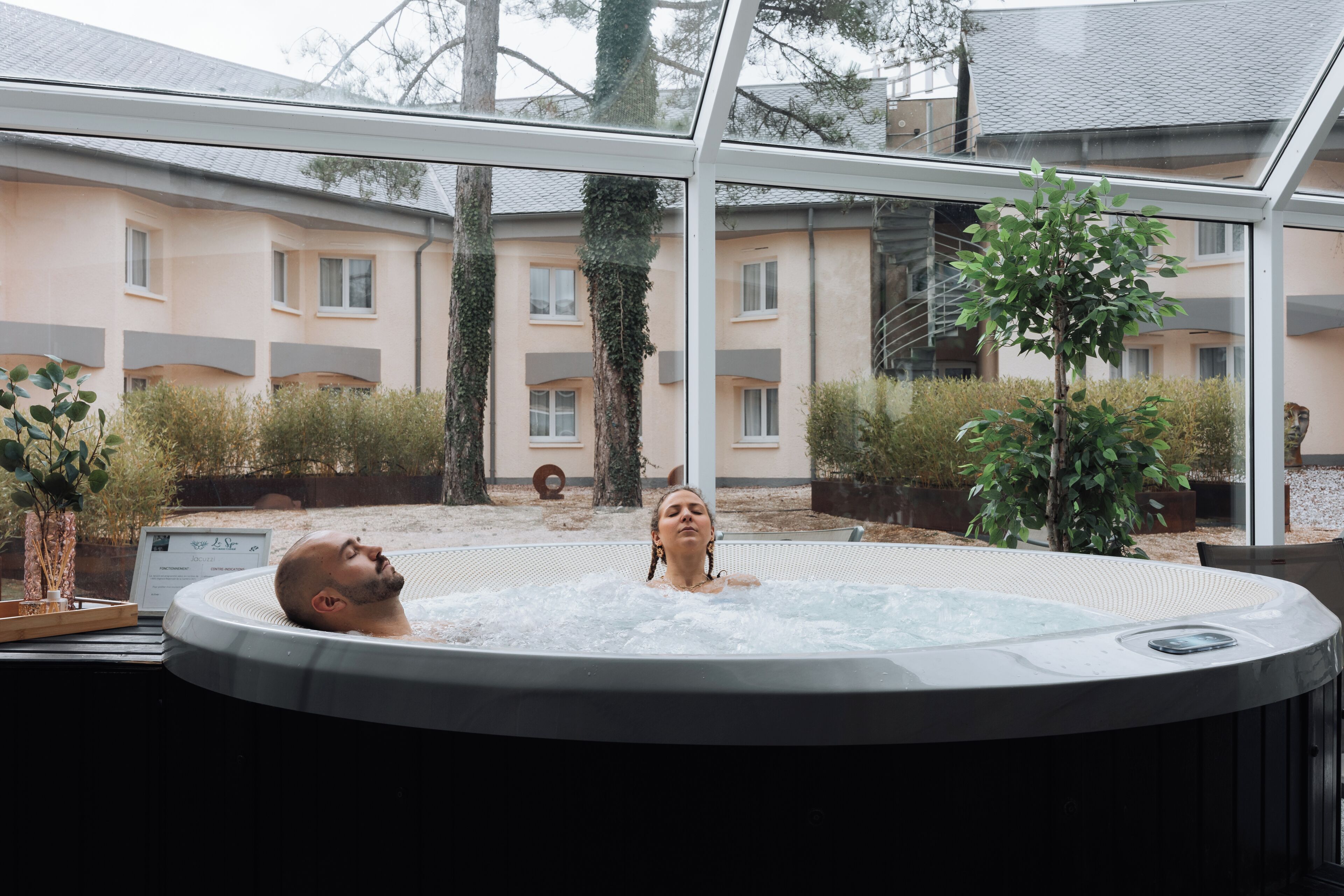 A hot tub with a glass enclosure, surrounded by outdoor plants and a building with multiple windows.