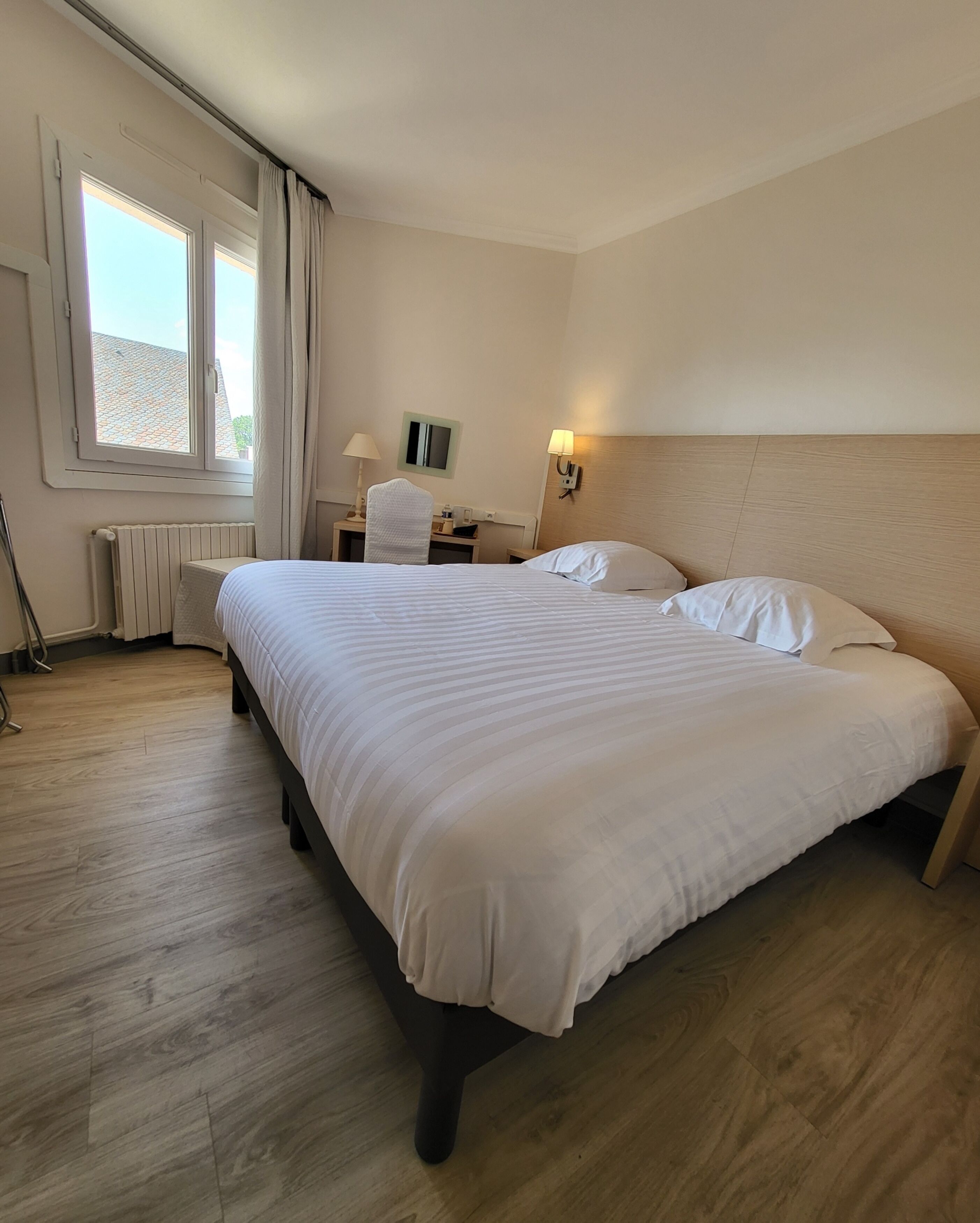 A hotel room with a large bed, a desk, a chair, a radiator, and a window with curtains.