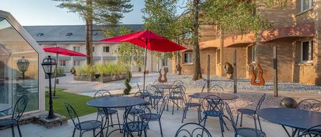 Un patio al aire libre con mesas y sillas, sombrillas rojas y un edificio con fachada de piedra al fondo.