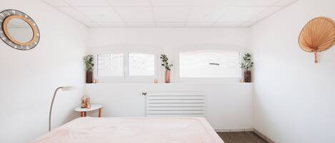 A minimalist bedroom with a bed, a radiator, a window with a view, a small table, and a floor lamp.