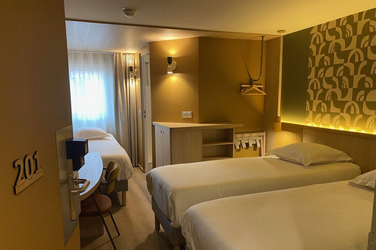 Ein Hotelzimmer mit zwei Betten, einem Schreibtisch und einem Stuhl. Ein Kleiderständer und eine kleine Kommode sind ebenfalls vorhanden.