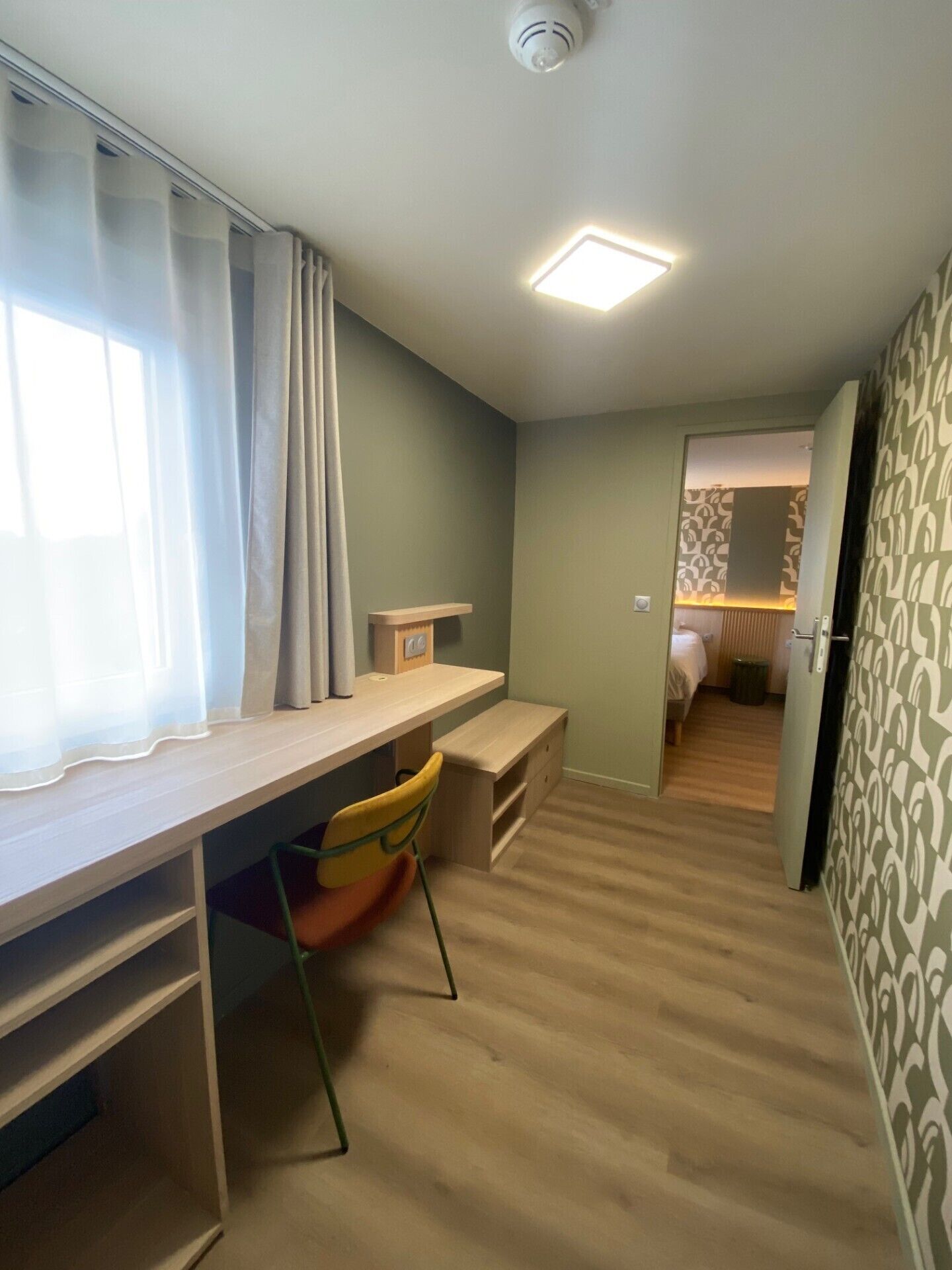 A compact room with a desk, chair, and a bed in the adjacent room.