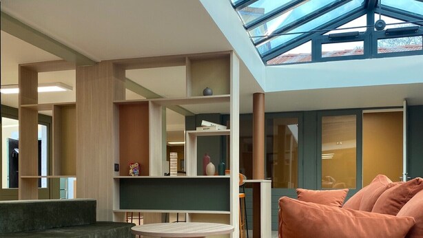 A modern interior with a high ceiling, a glass skylight, a seating area with a sofa and round tables, and a visible kitchen area. - Green Hôtels Poitiers (Poitiers)