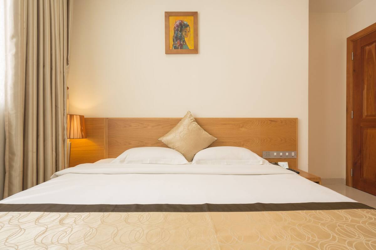 A hotel room with a large bed, a wooden headboard, a single bedside table, a lamp, and a painting on the wall.