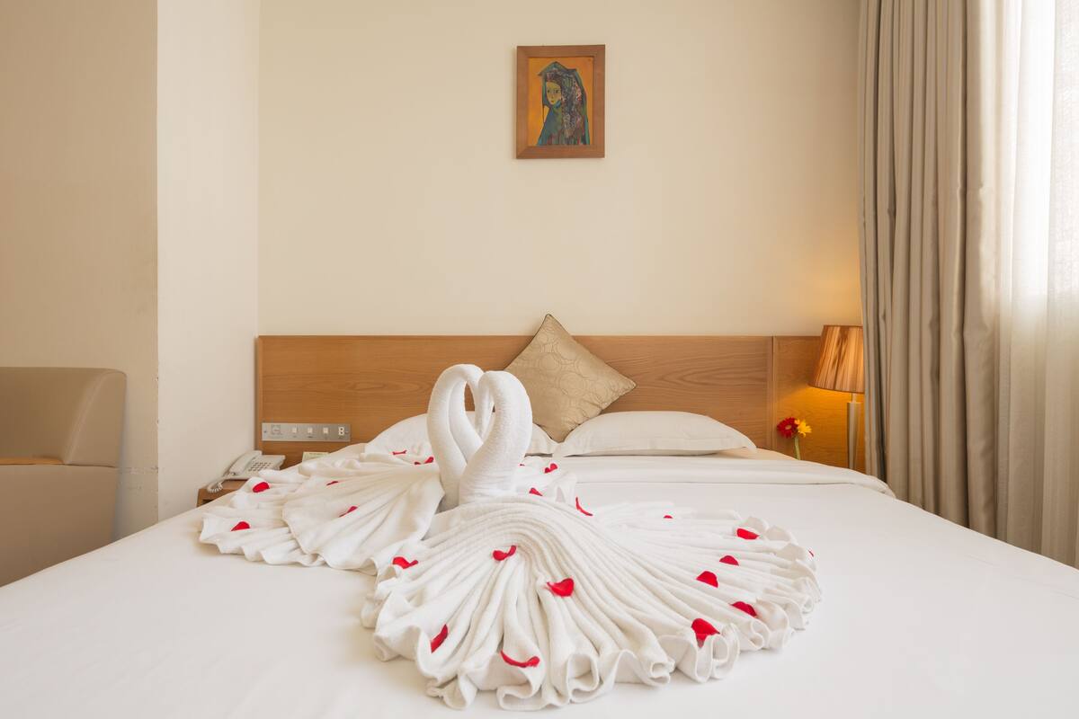 A neatly made bed with a towel arranged in the shape of two swans, a wooden headboard, a bedside lamp, and a painting on the wall.