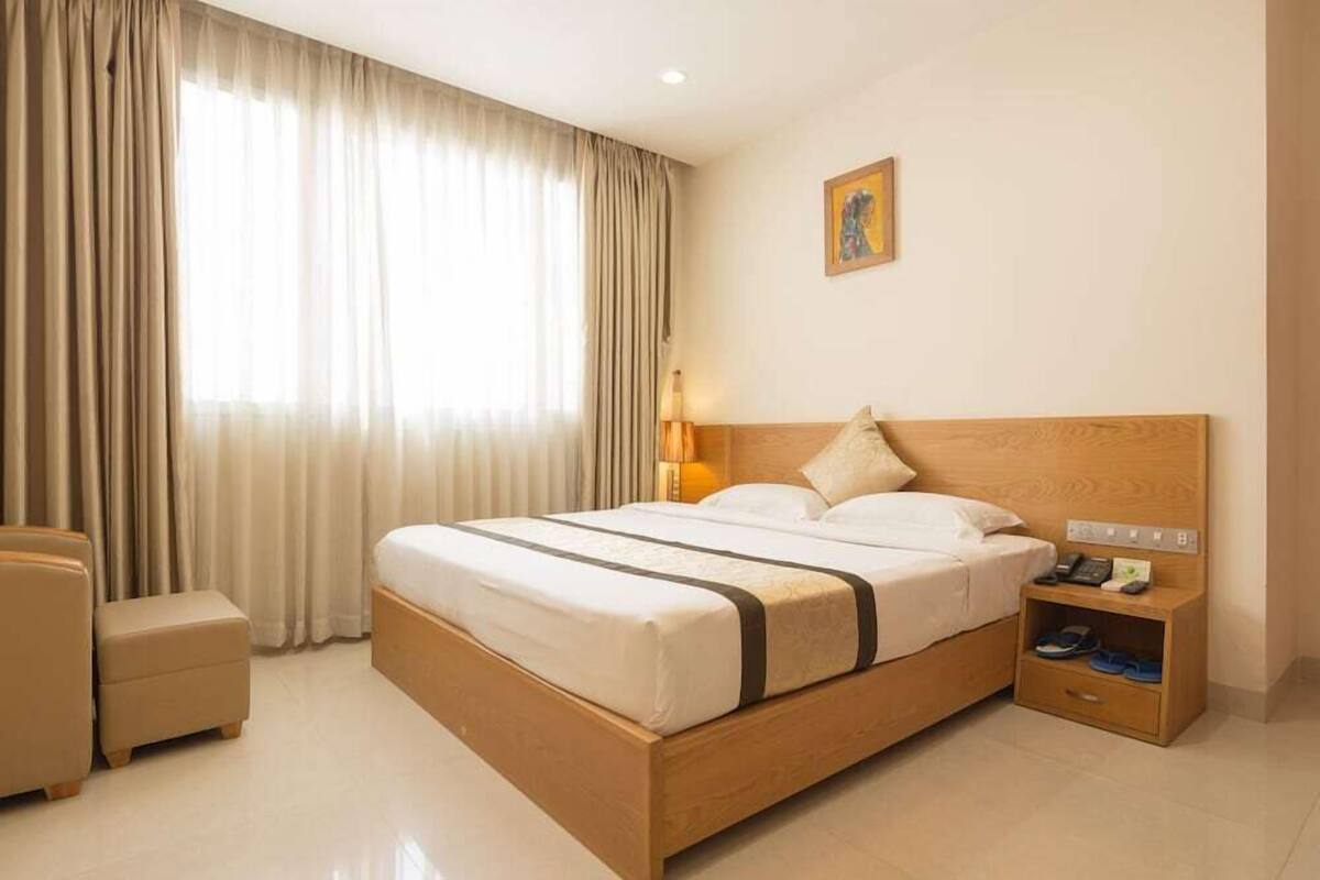 A hotel room with a wooden bed, a bedside table with a lamp, a chair, and a window with curtains.
