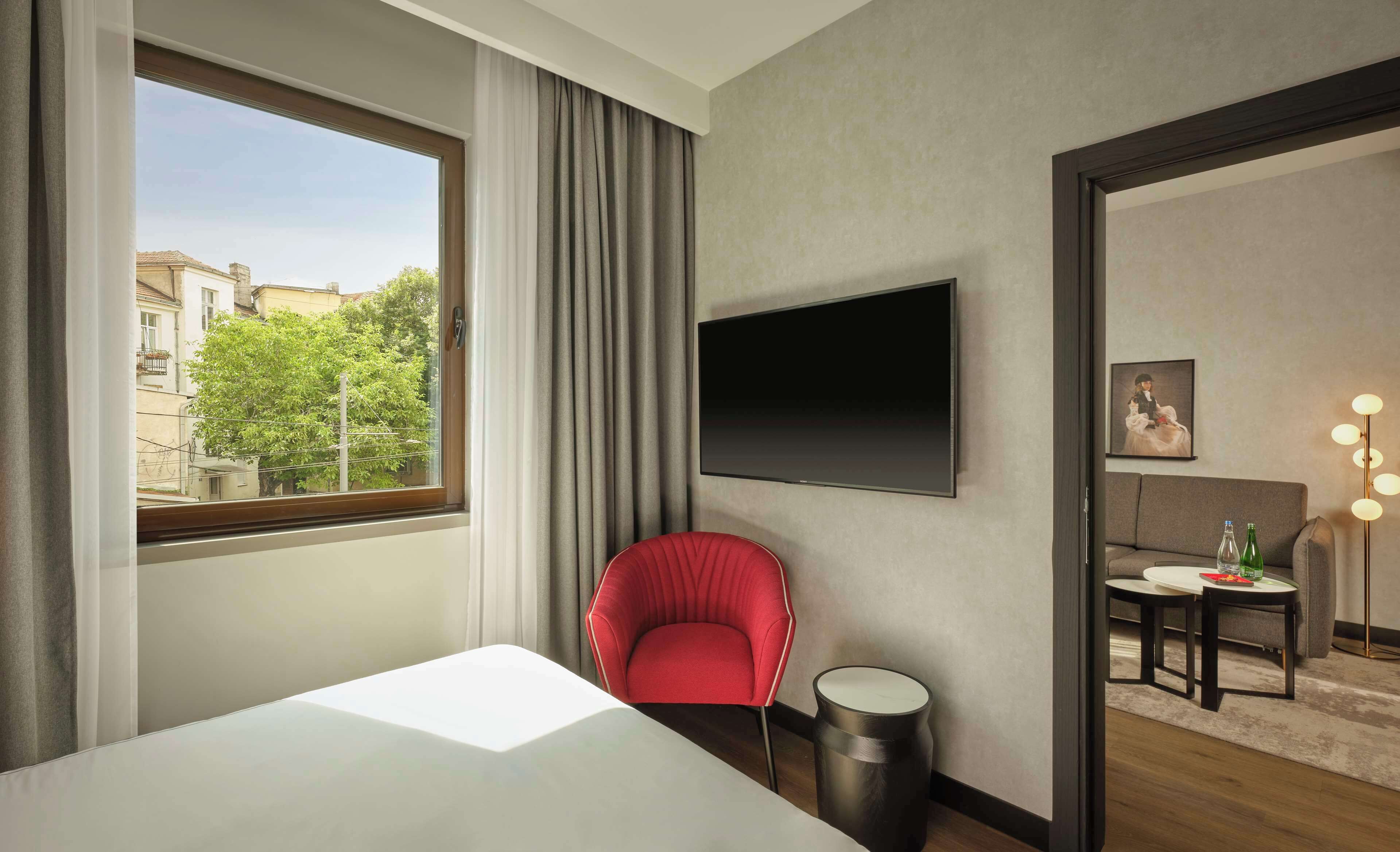 Habitación de hotel con cama, una silla roja, una mesa redonda con botellas, un televisor en la pared y vistas a edificios y árboles.