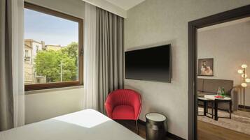 Habitación de hotel con cama, una silla roja, una mesa redonda con botellas, un televisor en la pared y vistas a edificios y árboles.