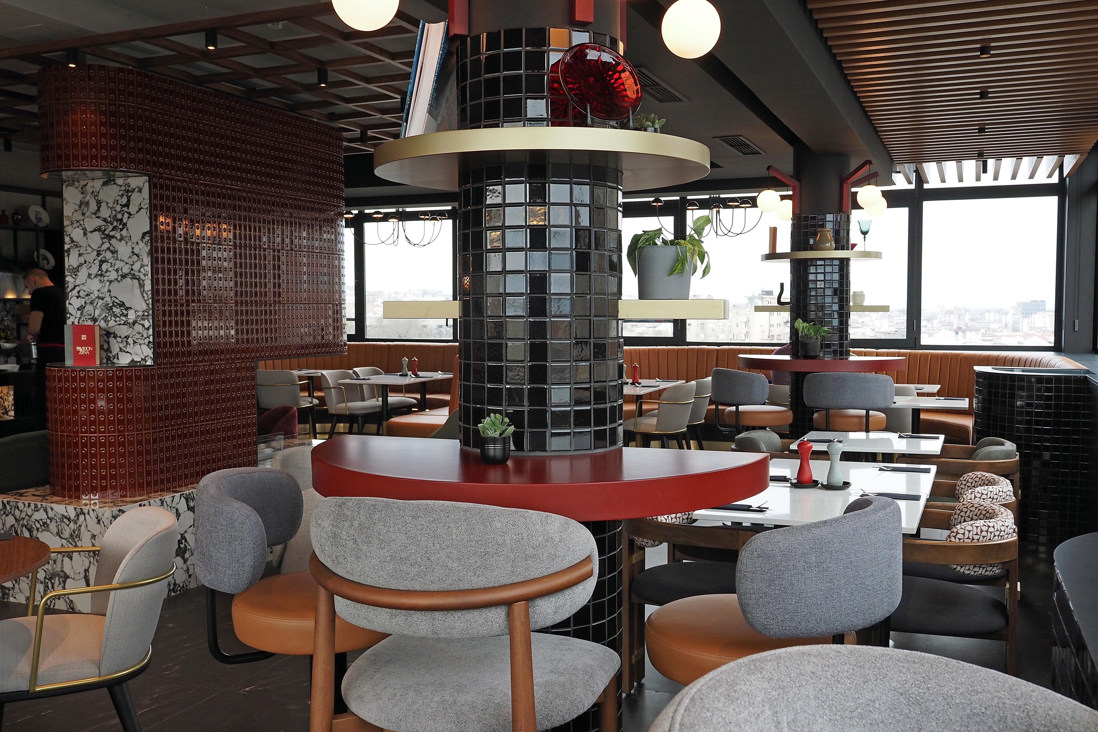 Un interior de restaurante moderno con mesas redondas, sillas tapizadas en gris y una columna decorativa central.