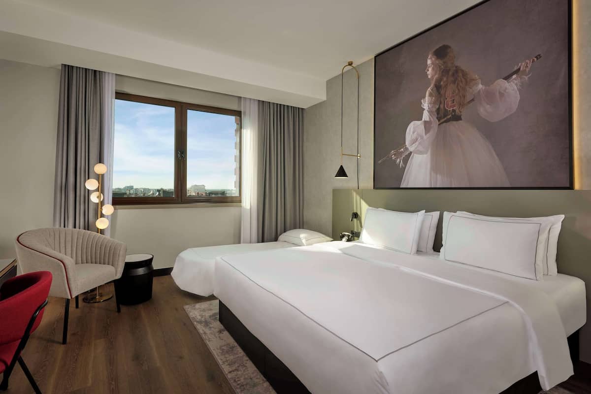 Une chambre d’hôtel avec un grand lit, un coin salon avec un fauteuil et une table, une fenêtre avec des rideaux et un tableau au mur.