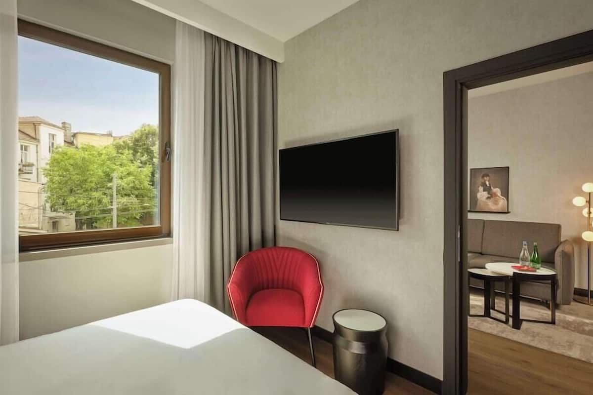 Une chambre d’hôtel avec un lit, une chaise rouge, une petite table ronde, un téléviseur fixé au mur et une vue sur les bâtiments et les arbres, visible par la fenêtre.