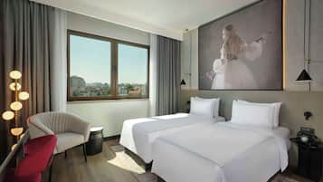 A hotel room with a large bed, a chair, a desk, and a view of the city through a large window.