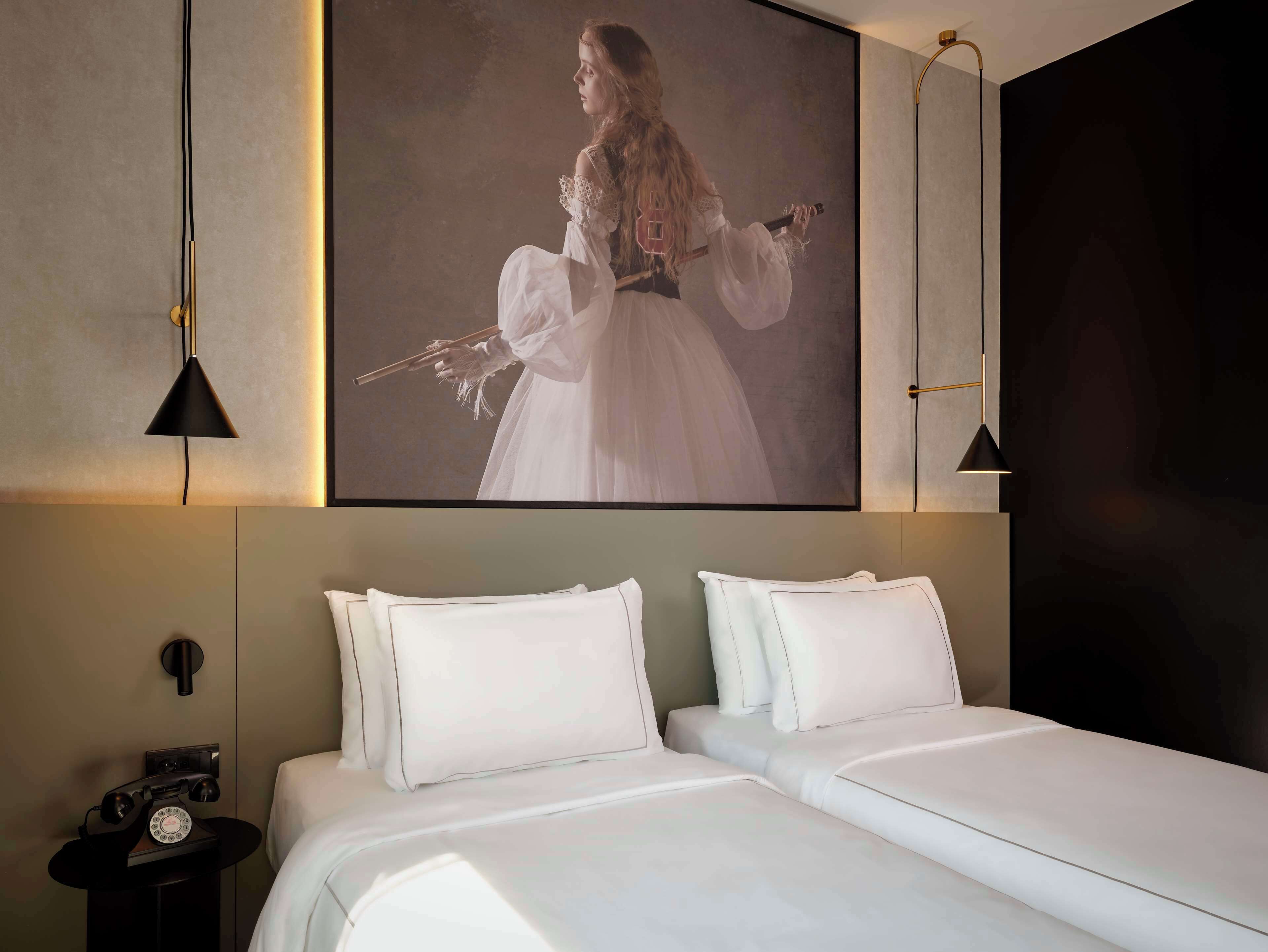 A hotel room with a large framed portrait, a bed with white linens, two bedside tables, and a classic telephone.