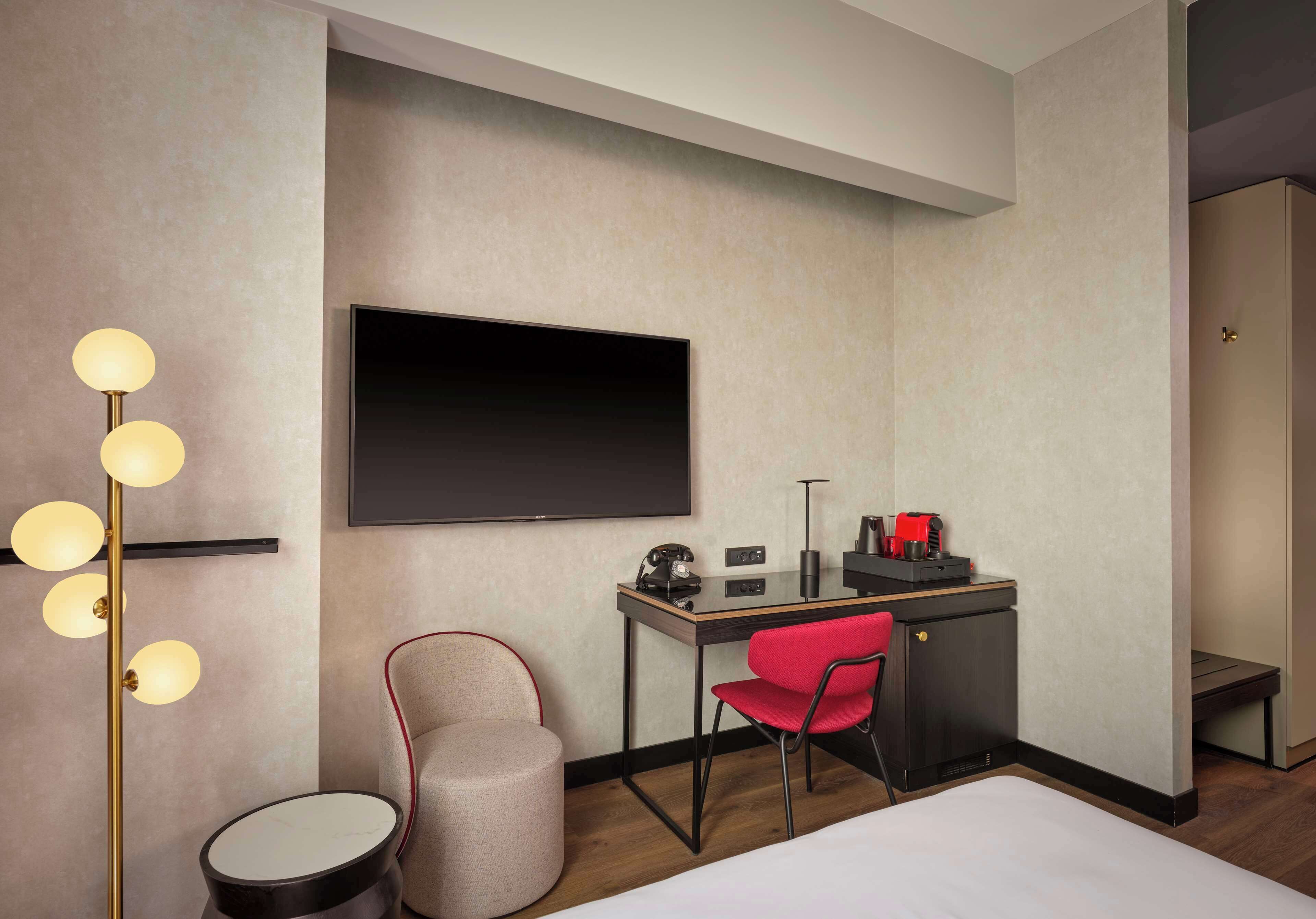 Una habitación de hotel moderna con una cama, un escritorio con silla, una mesa redonda, una silla roja, una lámpara de pie y un televisor de pantalla plana montado en la pared.