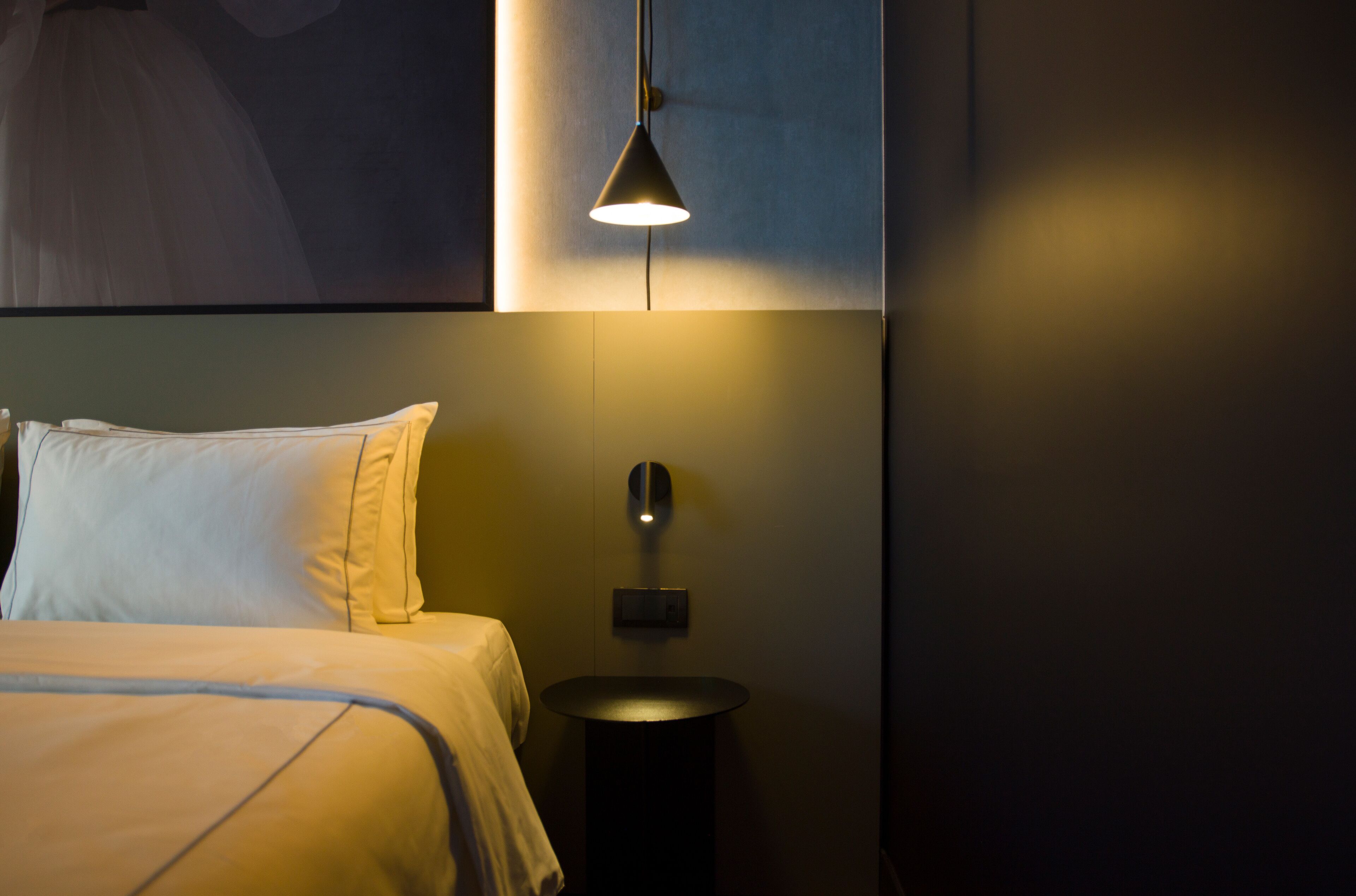 A neatly made bed with white linens, a bedside table with a lamp, and a wall-mounted light fixture.