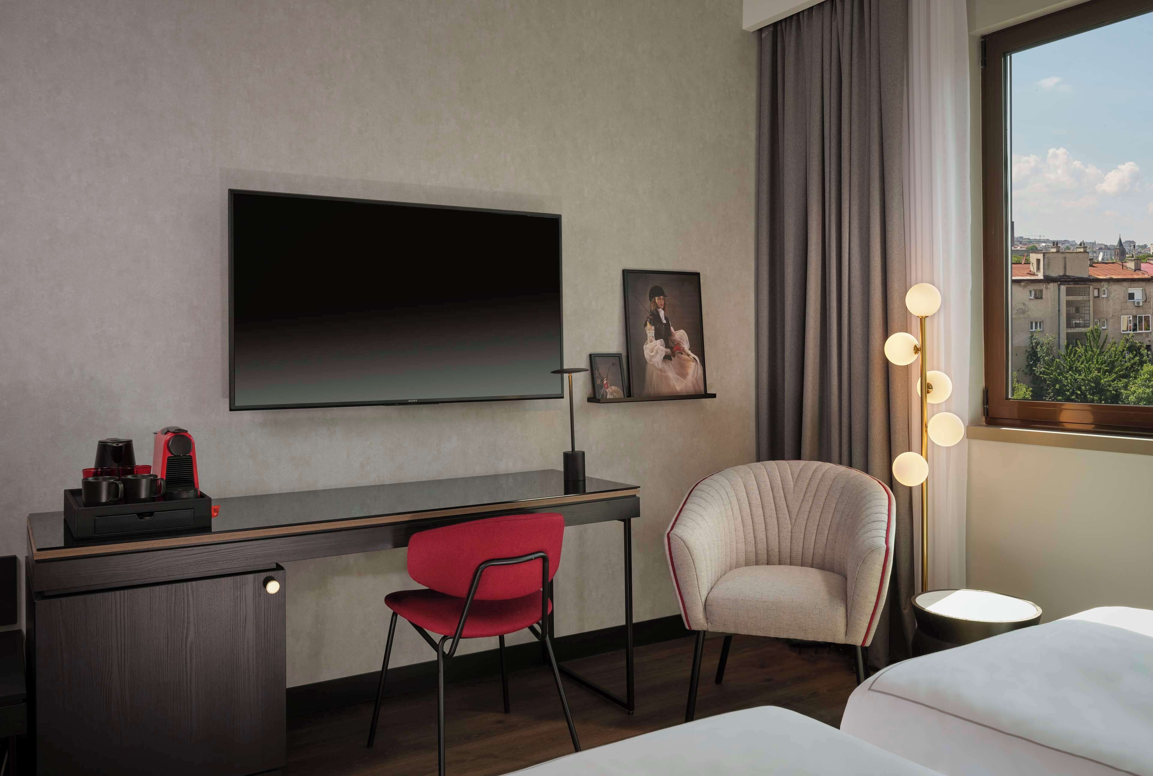 Ein modernes Hotelzimmer mit einem großen Flachbildfernseher an der Wand, einem Schreibtisch mit einem roten Stuhl, einem gepolsterten Sessel und einem Nachttisch mit einer Lampe.